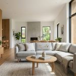 Interior ruang tamu bergaya Skandinavia yang modern, menampilkan sofa abu-abu, lantai kayu terang, jendela besar dengan cahaya alami, dan tanaman hias.